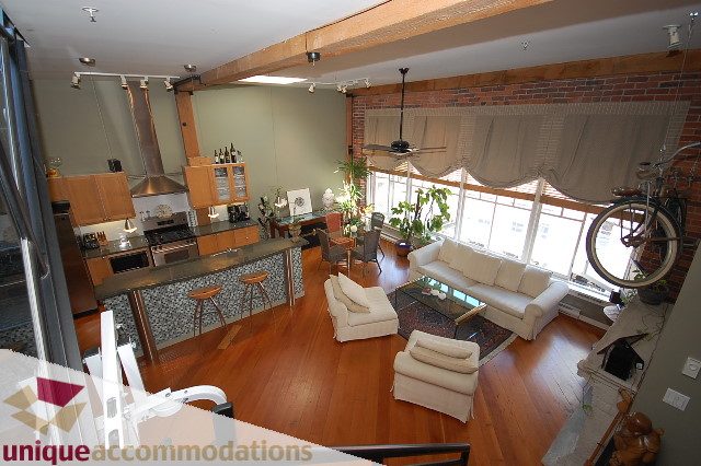 Bright kitchen with granite countertops and hardwood floors, open to a cozy living area with a brick wall and large windows.