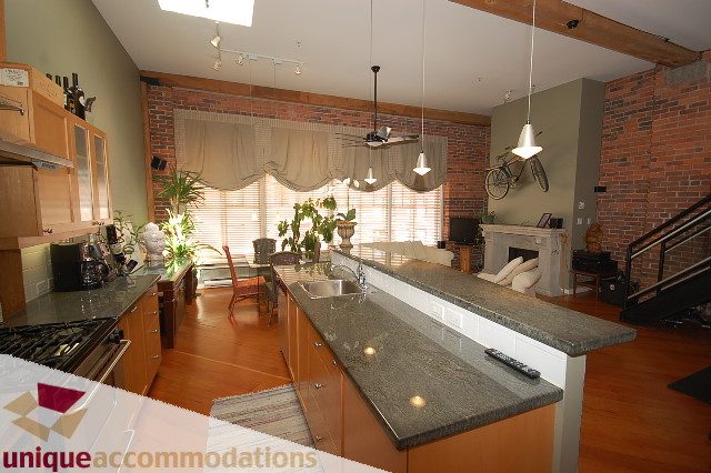 Bright kitchen with granite countertops and hardwood floors, featuring a cozy living area with brick walls and large windows.