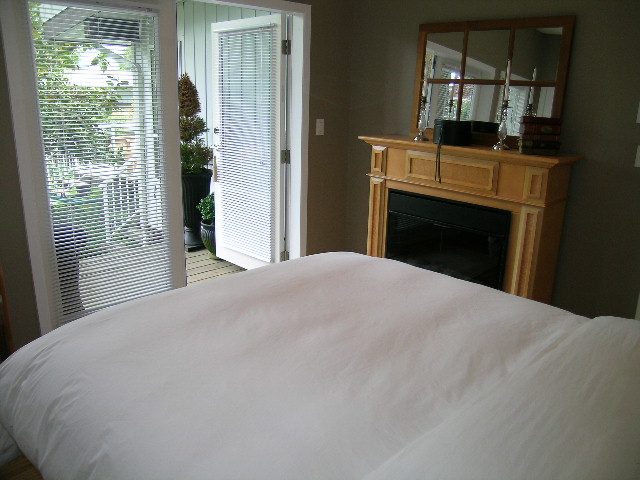Bright bedroom with white bedding, wooden fireplace, and sliding glass door leading to a patio. - 470 Ellis Street, North Vancouver