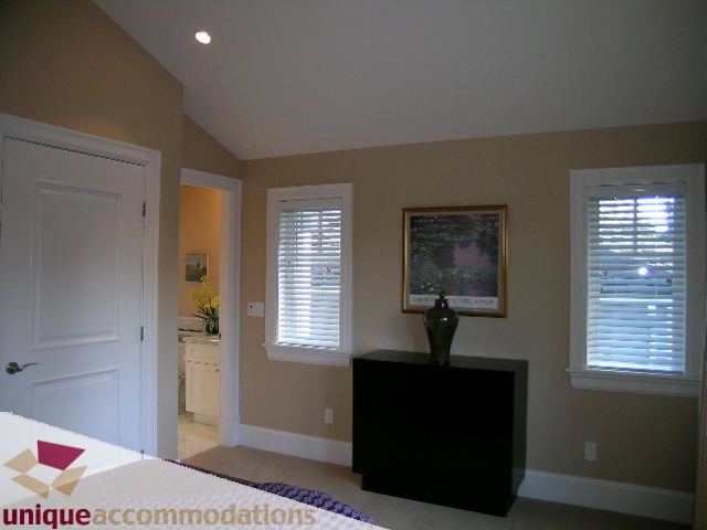 Bright bedroom with white blinds and a large window, featuring a black dresser and a mounted TV. - 1365 Kings Ave, West Vancouver