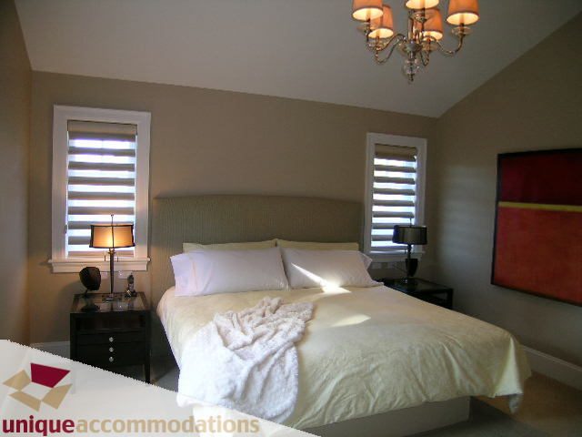 Furnished bedroom with plush bedding and elegant chandelier, featuring large windows with blinds. - 1365 Kings Ave, West Vancouver