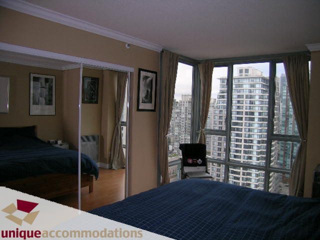 Bright bedroom with large windows offering a stunning city view, hardwood floors, and a neatly made bed.