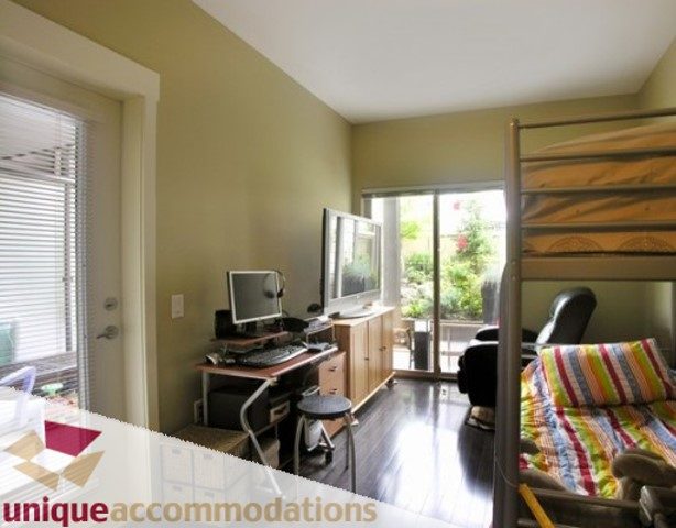 Bright living room with hardwood floors, a desk setup, and a large window offering a view of greenery. - 107 - 188 West 29th Street, North Vancouver