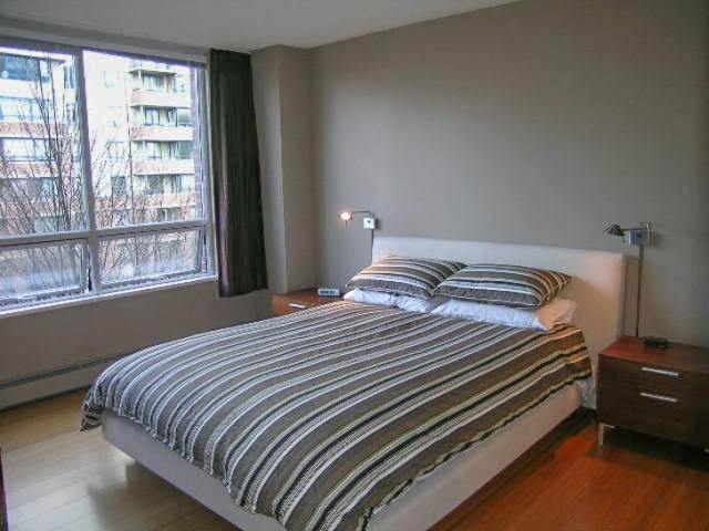 Spacious bedroom with hardwood floors, large windows offering city views, and neatly made beds with striped bedding.