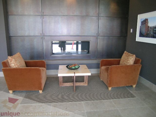 Furnished living room with modern fireplace, brown armchairs, and cityscape art. - 208 - 1833 Crowe St., Vancouver West Side