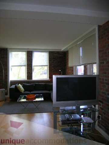 Furnished living room with hardwood floors and brick walls, featuring a glass coffee table and a black sofa.
