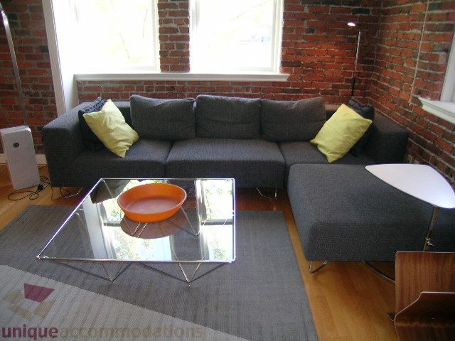Furnished living room with brick wall, modern sectional sofa, glass coffee table, hardwood floors, and natural light.