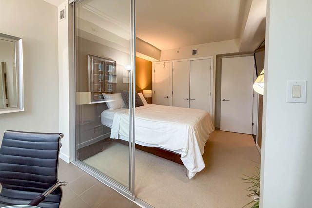Spacious 1-bedroom + den condo with a cozy bedroom featuring a neatly made bed, mirrored closet doors, and a modern chair.