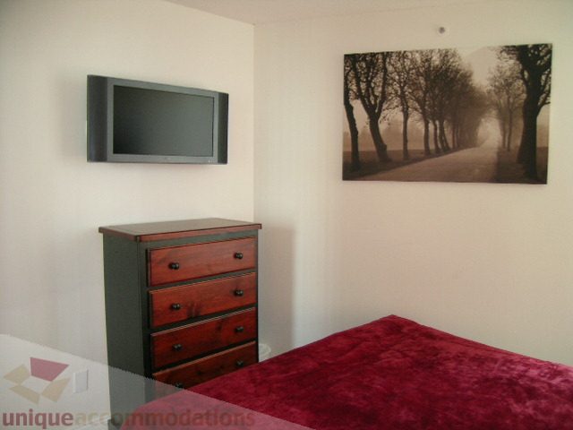 Furnished bedroom with a red bedspread, wooden dresser, and wall art of a tree-lined road. - 1403 - 555 Jervis Street, Vancouver Downtown