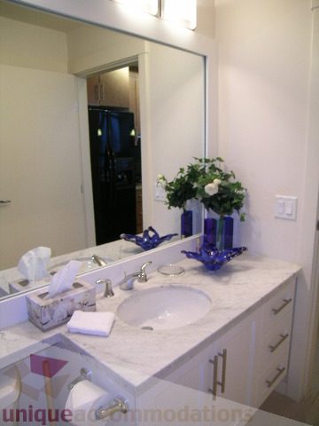 Furnished bathroom with marble countertops and a large mirror reflecting a kitchen area. - 3802 - 1189 Melville Street, Vancouver Downtown
