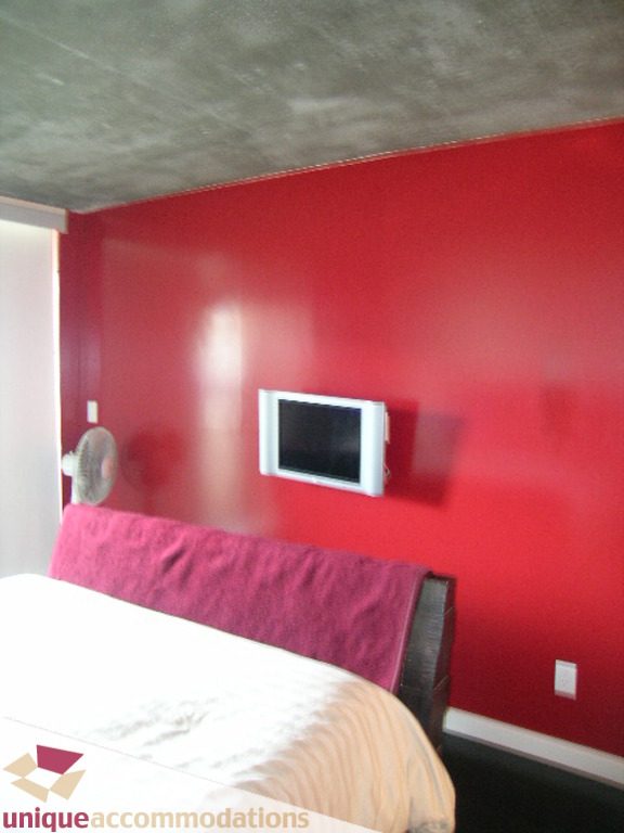 Spacious bedroom with red accent wall and mounted TV, featuring a cozy bed and concrete ceiling. - 705 - 1500 Howe street, Vancouver Downtown