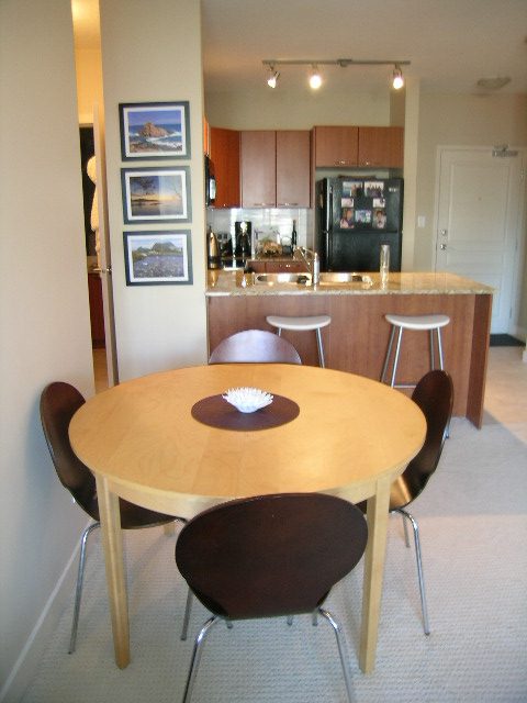 Bright dining area with wooden table and chairs, leading to a kitchen with granite countertops and bar stools.