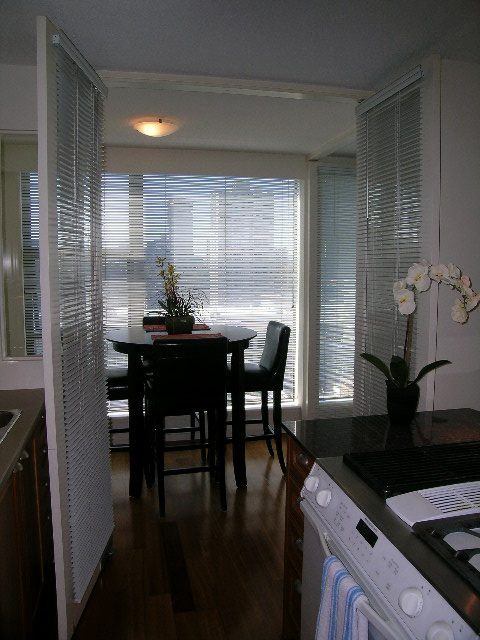 Bright kitchen with hardwood floors and a view of the dining area, featuring blinds and a potted plant.