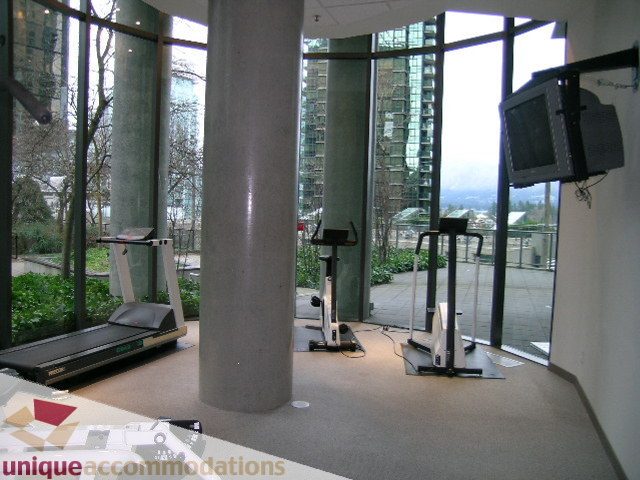 Spacious living room with large windows offering a stunning city view, featuring a treadmill and a flat-screen TV.