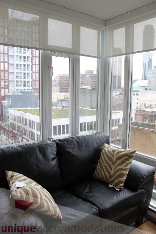 Furnished living room with a black leather couch, city view, and large windows. - 705 - 565 Smithe Street, Vancouver Downtown