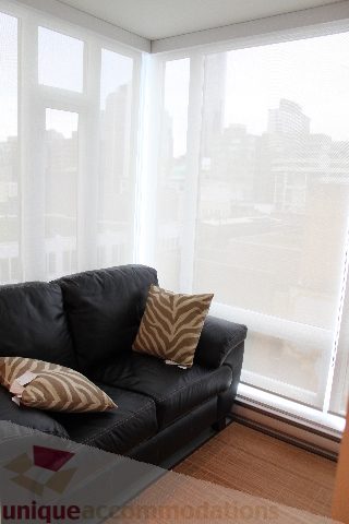 Furnished living room with a black leather couch, city view, and sheer curtains. - 705 - 565 Smithe Street, Vancouver Downtown