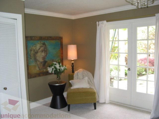 Spacious living room with a plush green chair, a painting of a woman, and a large bay window with white curtains.