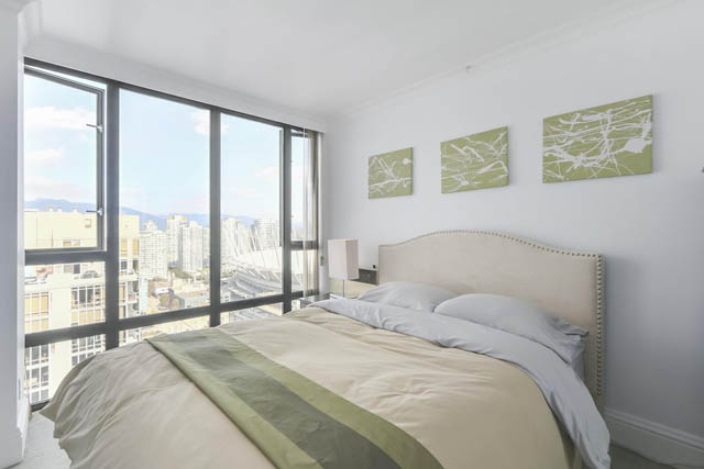 Bright bedroom with large windows offering city views, modern artwork on walls, and neatly made beds.