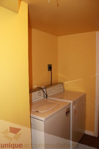 Small laundry room with yellow walls, white washer and dryer, and a small counter space. - 2156 Cortell Ave, North Vancouver