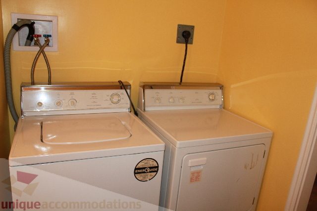 Laundry room with stacked washer and dryer, yellow walls, and visible utility connections. - 2156 Cortell Ave, North Vancouver
