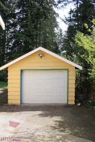 Bright garage with yellow siding and white door, surrounded by trees. - 2156 Cortell Ave, North Vancouver
