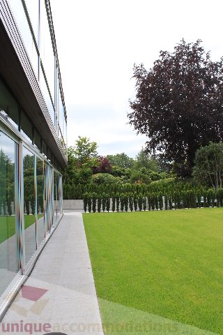 Furnished backyard with green lawn and trees, modern glass building in background. - 6012 Newton Wynd, Vancouver West Side