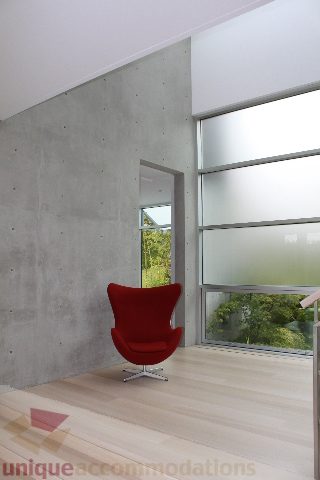 Bright living room with concrete wall, red chair, and large windows showing greenery. - 6012 Newton Wynd, Vancouver West Side