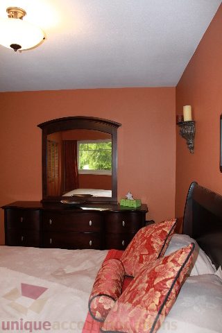 Furnished bedroom with a wooden dresser, red patterned pillows, and a view of greenery outside the window. - 2730 Ailsa Crescent, North Vancouver