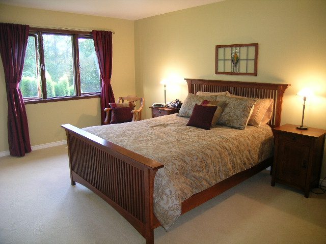 Spacious bedroom with wooden sleigh bed, patterned bedding, and large window with red curtains. - 5753 Westport Road, West Vancouver