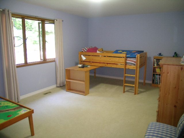 Bright bedroom with wooden bunk bed, desk, and bookshelf, light blue walls, and large window with curtains. - 5753 Westport Road, West Vancouver