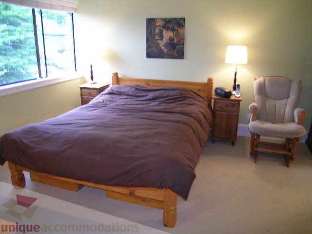 Spacious bedroom with wooden bed frame, neutral decor, and a cozy armchair. - 405 - 4001 Mount Seymour Pkwy, North Vancouver