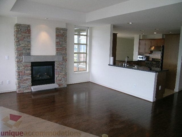 Open-concept living room with stone fireplace and hardwood floors, leading to a modern kitchen with granite countertops.