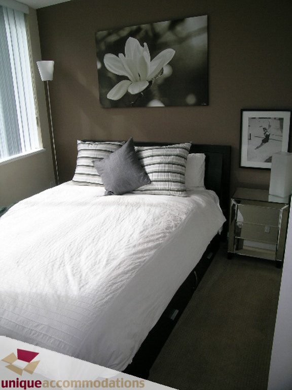 Furnished bedroom with white bedding, black and white artwork, and a nightstand with a lamp. - 316 - 1082 Seymour, Vancouver Downtown