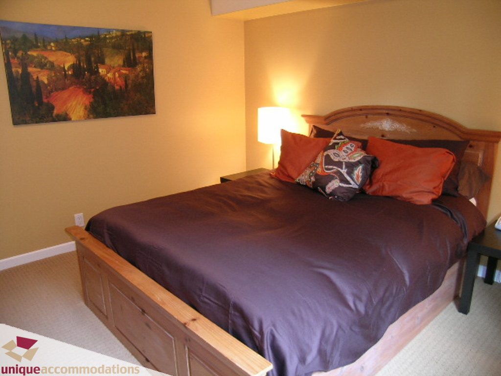 Furnished bedroom with wooden bed, purple bedding, and a warm lamp. - 1385 East 15th Street, North Vancouver