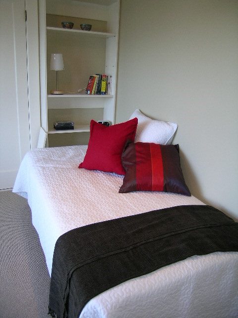 Bright bedroom with neatly made bed, red and brown pillows, and a small shelf with books and decor. - 4663 Bellevue Drive, Vancouver West Side