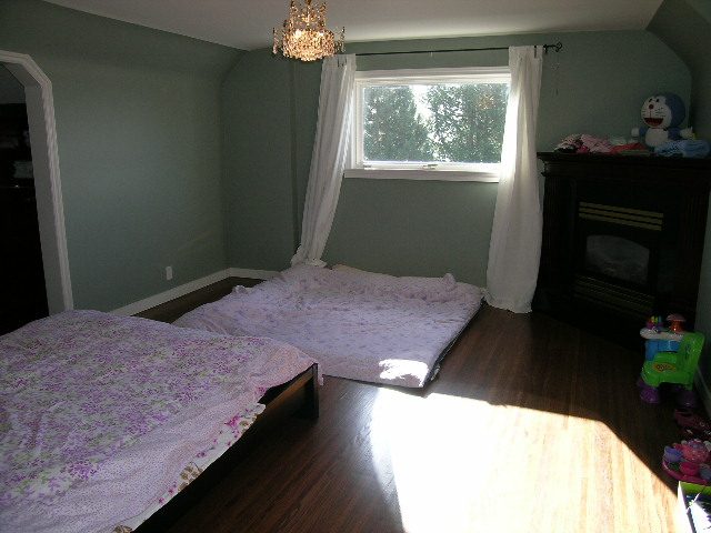 Spacious bedroom with hardwood floors, floral bedspread, and a large window with white curtains. - 1875 26th Street, West Vancouver
