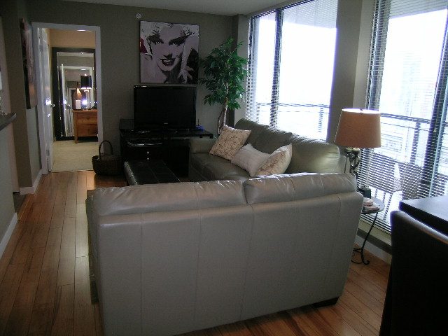 Spacious living room with hardwood floors, a large window offering a city view, and a comfortable leather sofa.