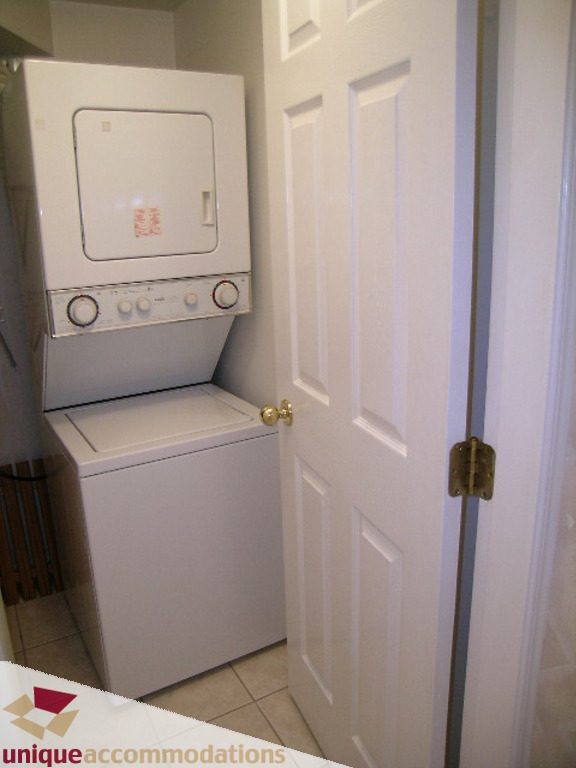 Compact laundry room with stacked washer and dryer, white door, and tiled floor. - 1339 West 7th Avenue, Vancouver West Side