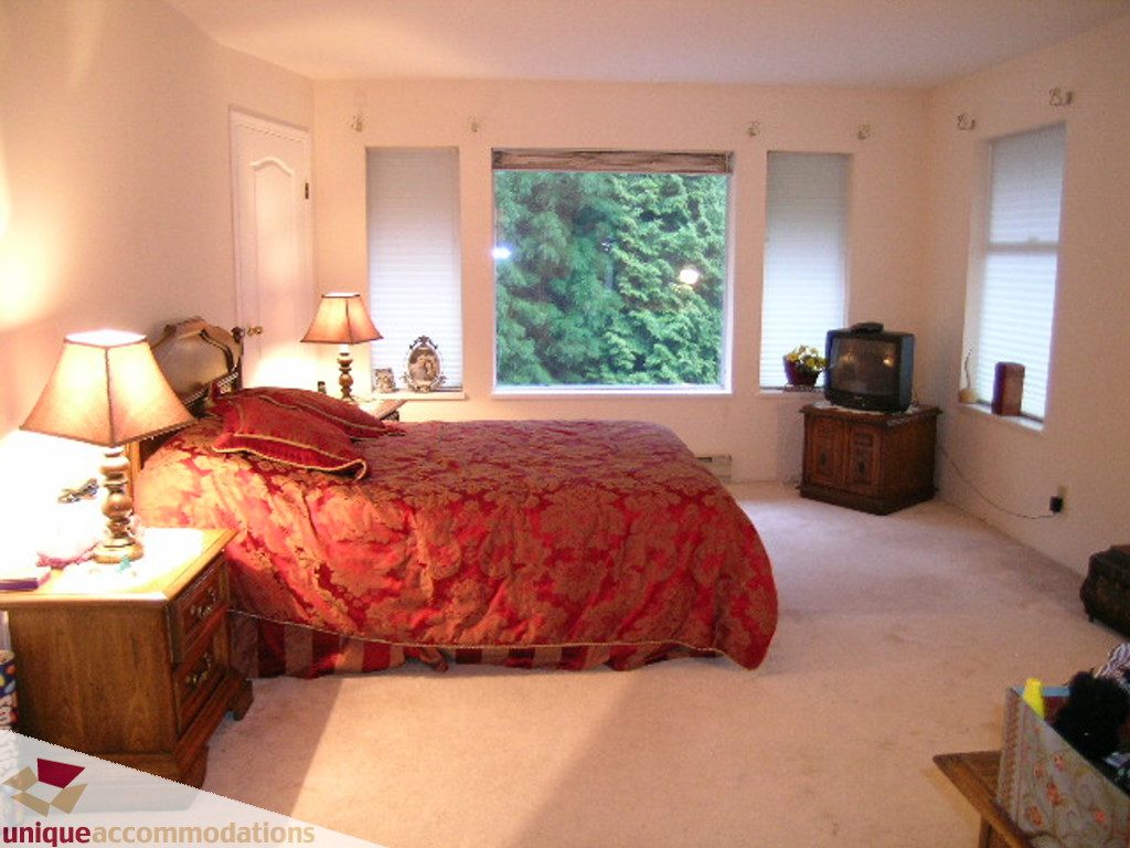 Bright bedroom with red patterned bedspread, large window view, and wooden nightstands. - 5735 Nancy Green way, North Vancouver