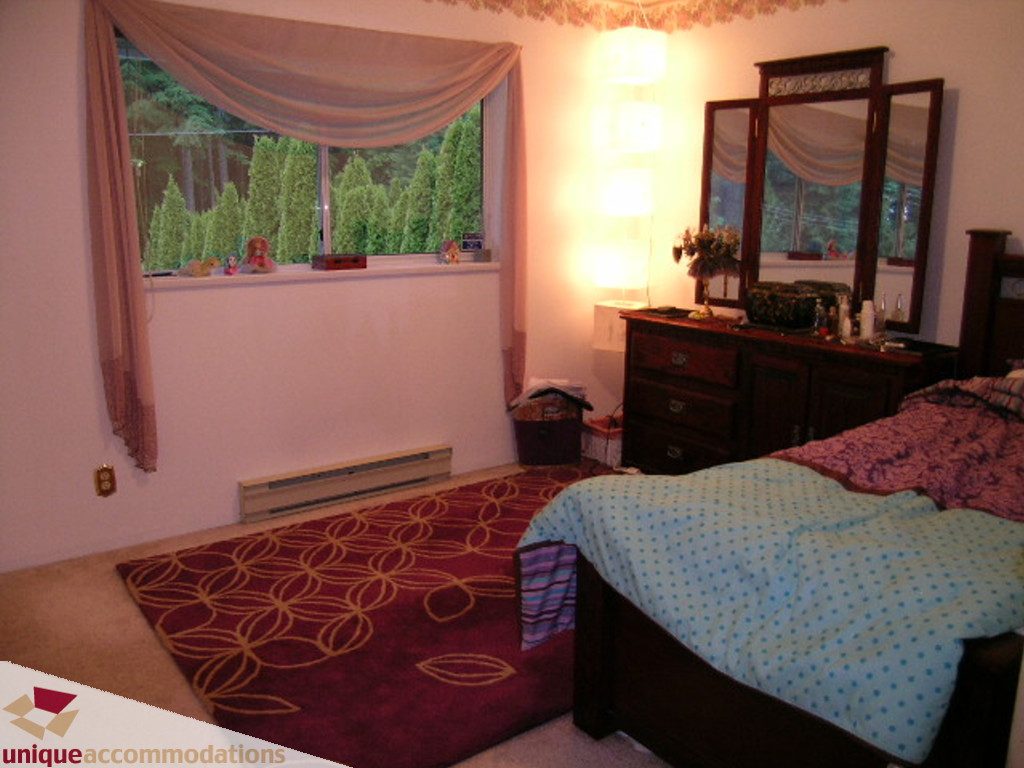 Furnished bedroom with a large window, floral rug, and dresser with mirror. - 5735 Nancy Green way, North Vancouver