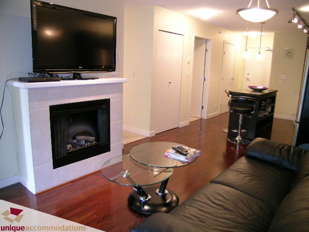 Furnished living room with hardwood floors, glass coffee table, and a flat-screen TV above a fireplace. - 305 - 538 Smithe St, Vancouver Downtown