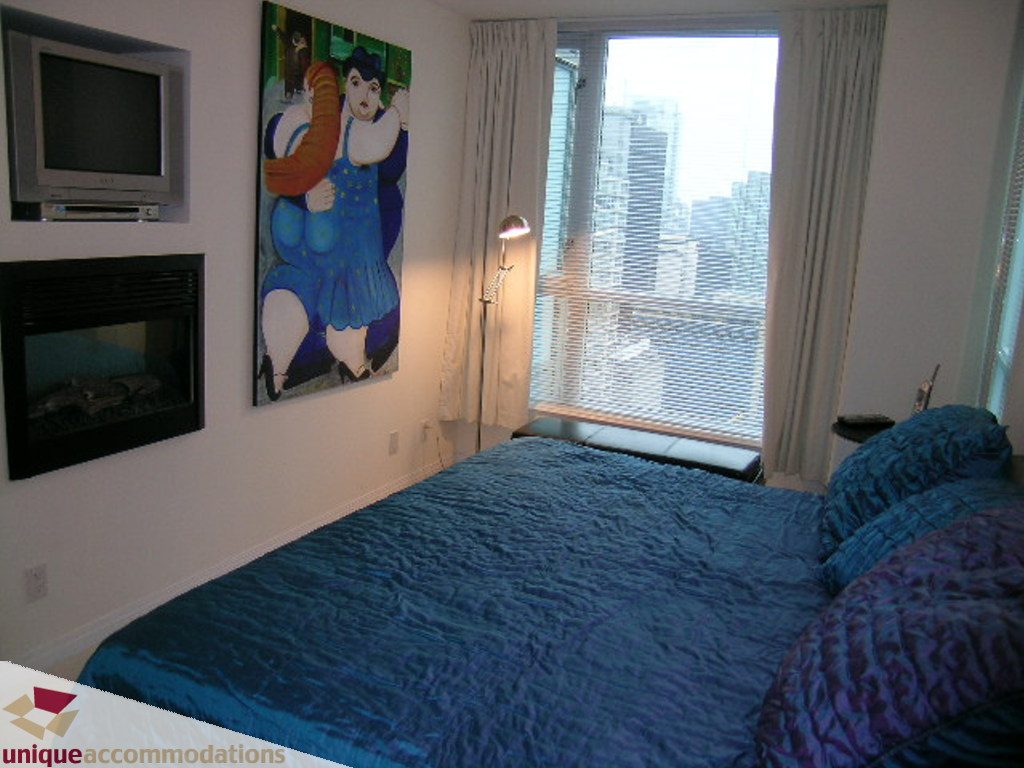 Furnished bedroom with blue bedding and city view through large window. - 2907 - 1328 W Pender St, Vancouver Downtown