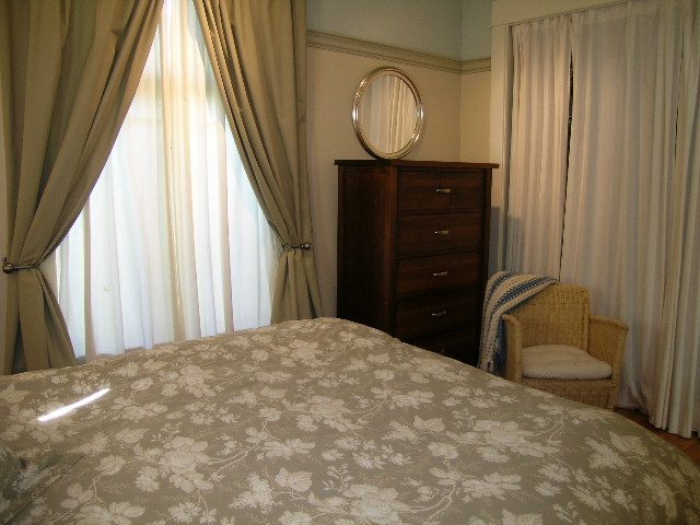 Furnished bedroom with floral bedspread, wooden dresser, and large window with curtains. - 3602 Fromme Rd, North Vancouver