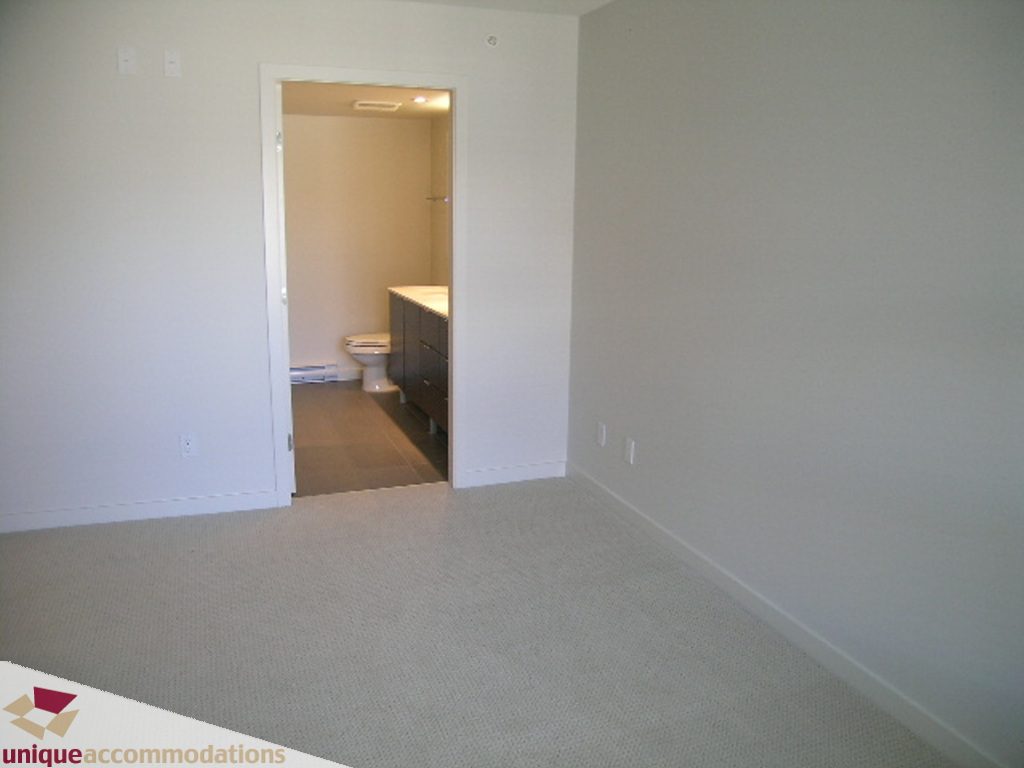 Bright living room with beige carpet, leading to a bathroom with white fixtures and a countertop. - 510 - 1677 Lloyd Avenue, North Vancouver