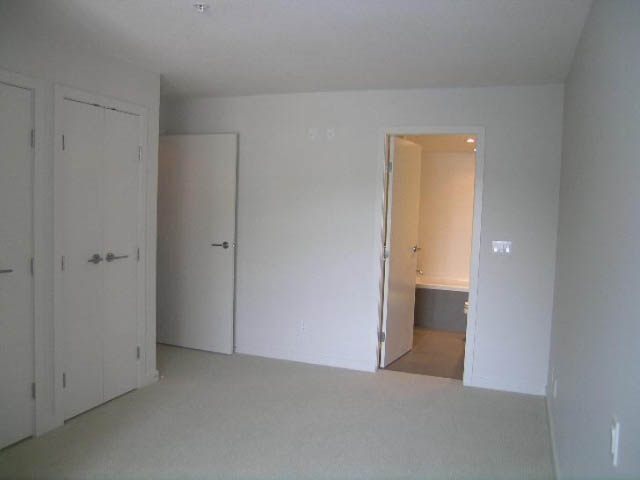 Bright living room with carpeted floor and white walls, leading to another room. - 302 - 1677 Lloyd Avenue, North Vancouver