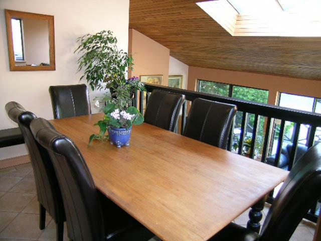 Dining area with wooden table, black leather chairs, and large windows showing greenery. - 1077 Blue Grouse Way, North Vancouver