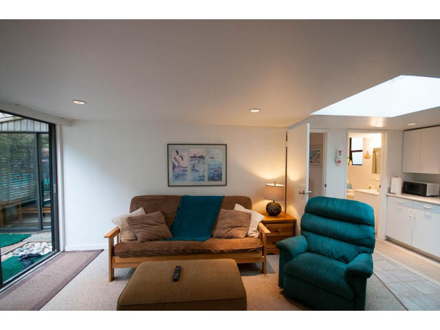 Bright living room with teal armchair and wooden coffee table, kitchen in background. - Cottage - 1395  3rd Street, West Vancouver