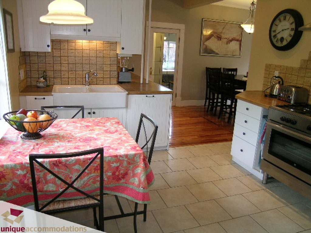 Bright kitchen with white cabinetry, hardwood floors, and a cozy dining area, featuring a floral tablecloth and a clock...