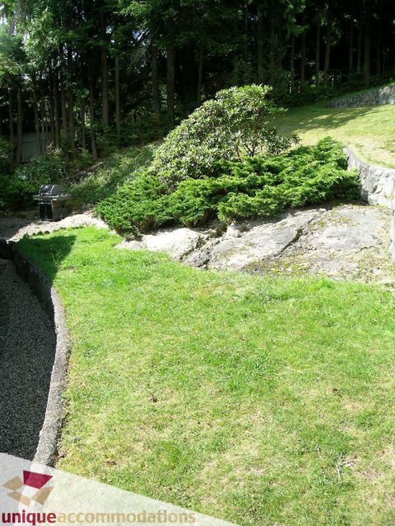 Lush green lawn with a large bush and rocky area, surrounded by trees. - 68 Bonnymuir Drive, West Vancouver