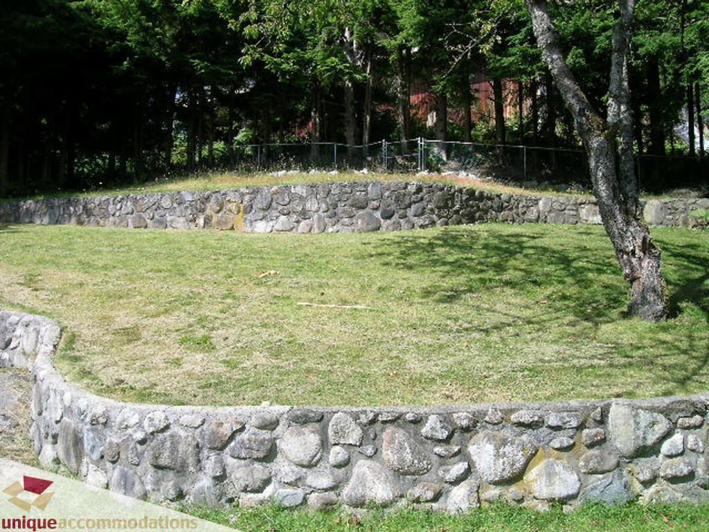 Fenced backyard with stone walls and grassy area, surrounded by trees. - 68 Bonnymuir Drive, West Vancouver